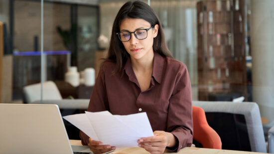 Focused latin hispanic business woman working on laptop computer reading financial document trader report in office. Thinking accountant entrepreneur manager businesswoman doing paperwork using pc